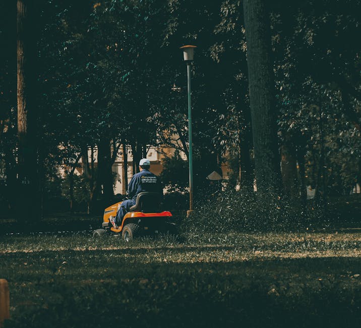 A person wearing a dark long-sleeved shirt and a grey hat is trimming a well-maintained hedge with long-handled hedge shears in a lush garden. The hedge, composed of dense, small green leaves, runs ho