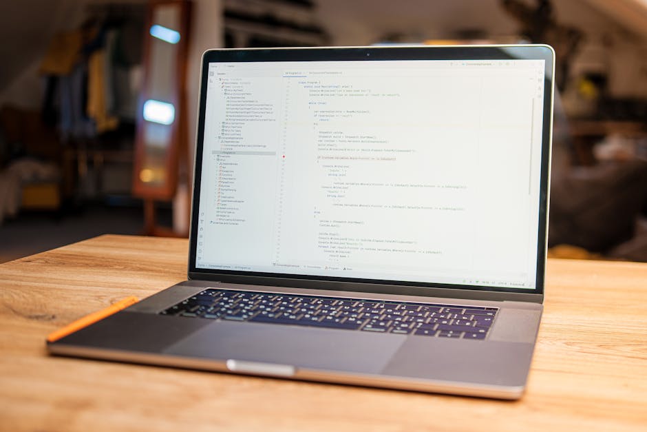 A close-up view of a laptop placed on a wooden table in a well-lit indoor space, displaying lines of code on its screen amidst a blurred background featuring furniture and warm lighting. The laptop ha